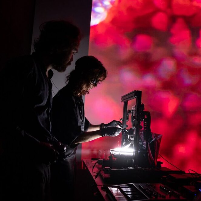 Baudouin Saintyves et Séverine Atis au festival Ars Electronica 2024 avec Water Organoids. On les voit en contre-jour en train de réaliser les expériences qui sont projetées sur écran, en fond dans les tons rouges